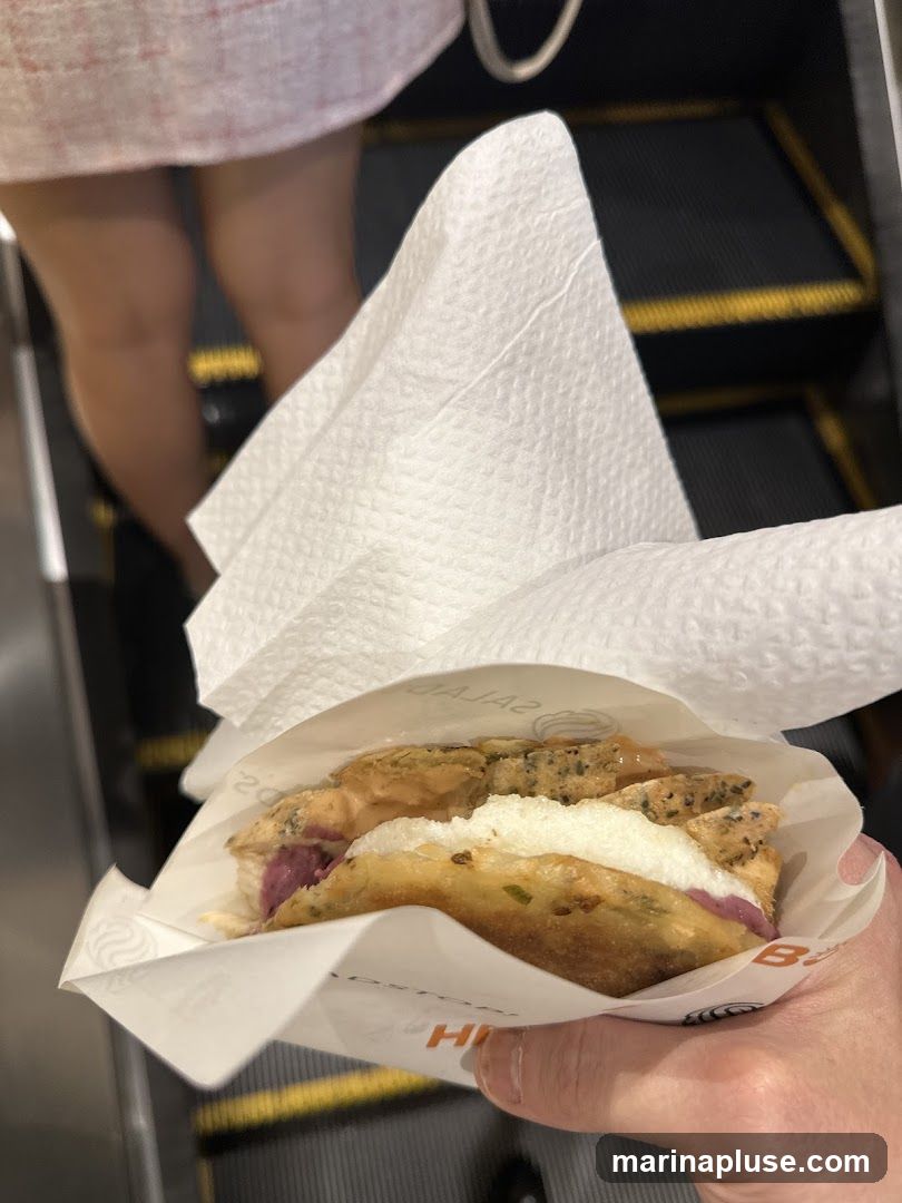 A person holds a sandwich wrapped in paper on an escalator. The sandwich appears to have layers of fried dough, white filling, and purple ingredients. The paper wrapping has visible branding, including the words "SALADS" and "HI". The background shows the steps of a dark escalator with yellow safety lines.