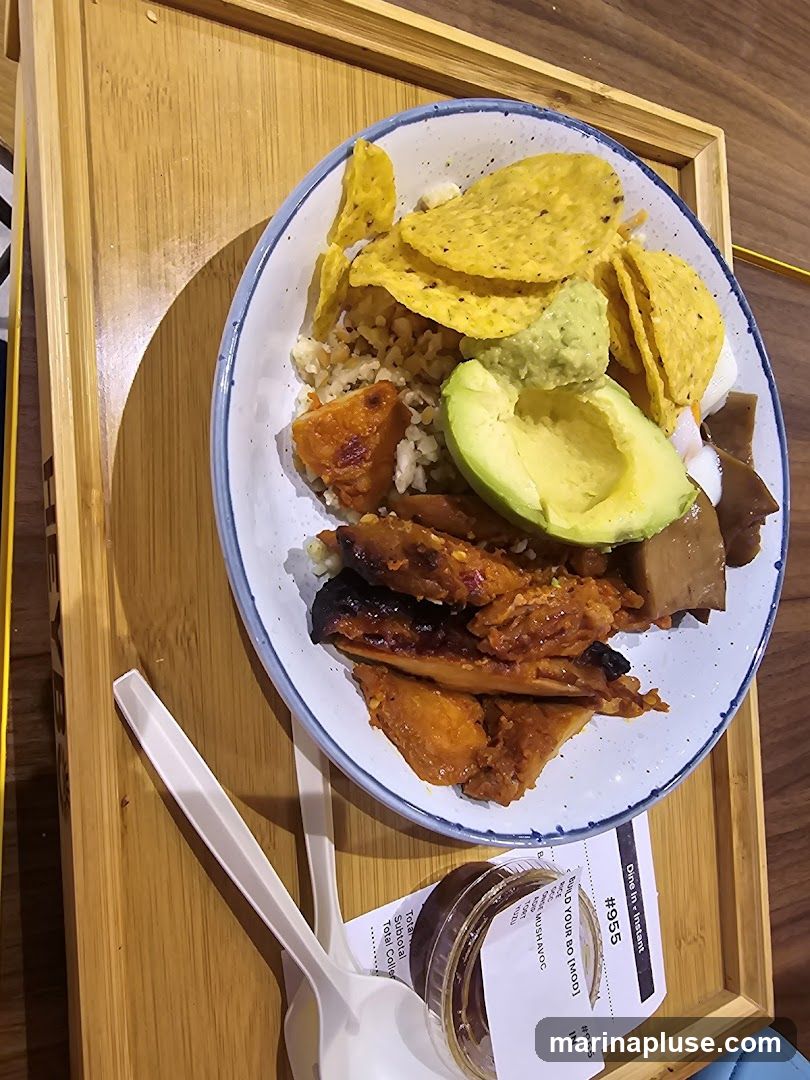 A plate of food containing rice, sweet potato, grilled chicken or tofu, guacamole, avocado slices, and tortilla chips. The meal is served on a bamboo tray with plastic cutlery and a small container of sauce. A receipt is visible under the plate.