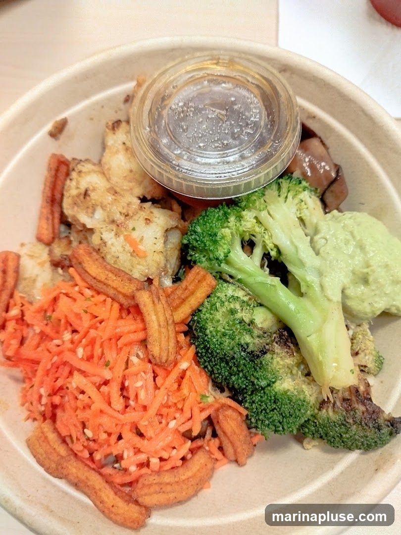 A close-up shot of a bowl of food. The bowl contains shredded carrots, fried pieces of fish, broccoli florets, and a dollop of green sauce. There are also some fried churro-like pieces scattered throughout the bowl. A small plastic container with a dark liquid is placed on top of the food.