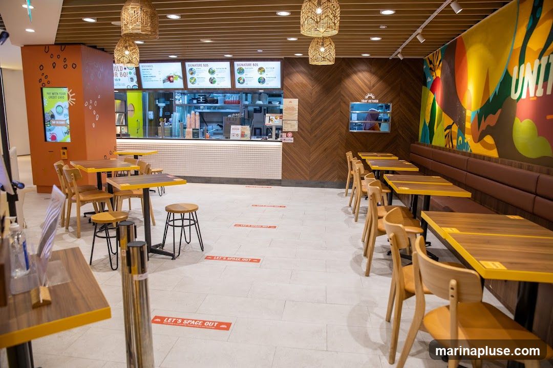 The image shows the interior of a fast-casual restaurant. There are several tables and chairs arranged for dining. The counter area is visible in the background with menu boards above it. A large, colorful mural adorns one wall. Social distancing markers are visible on the floor.