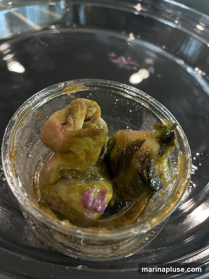 A close-up shot of pickled Brussels sprouts in a small glass bowl. The sprouts are green with some purple discoloration and are submerged in a light brown liquid. The bowl is placed on a dark, reflective surface.