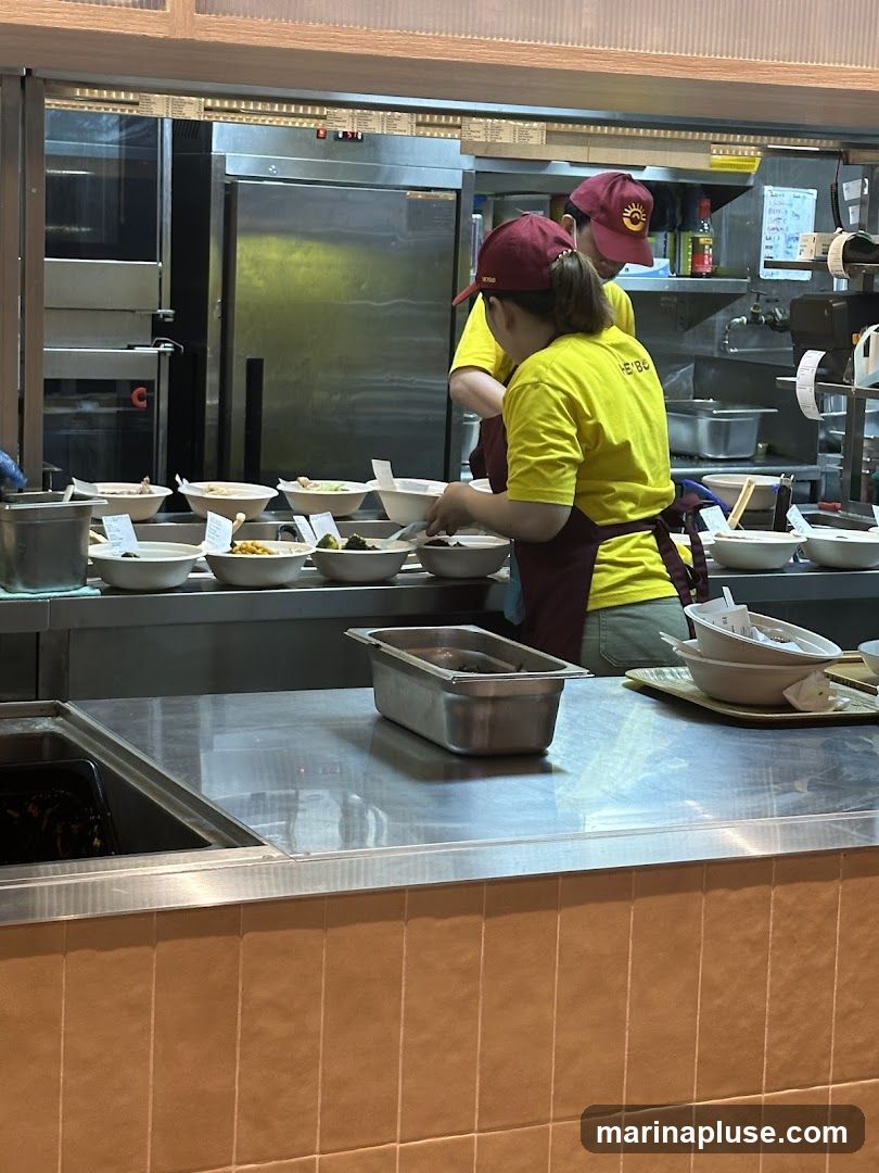 Two employees in yellow shirts and maroon caps are preparing food behind a counter. The counter is made of stainless steel and has several bowls of food on it. There are also ovens and other kitchen equipment in the background.