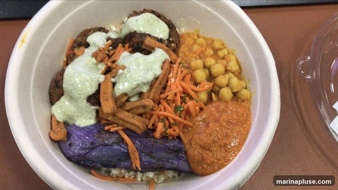 A close-up overhead view of a take-out bowl filled with a Middle Eastern-inspired meal. The bowl contains rice, a piece of purple eggplant, falafel patties drizzled with a green sauce, shredded carrots, crispy fried pieces, chickpeas in a sauce, and a dollop of orange sauce. A clear plastic lid is partially visible to the right.