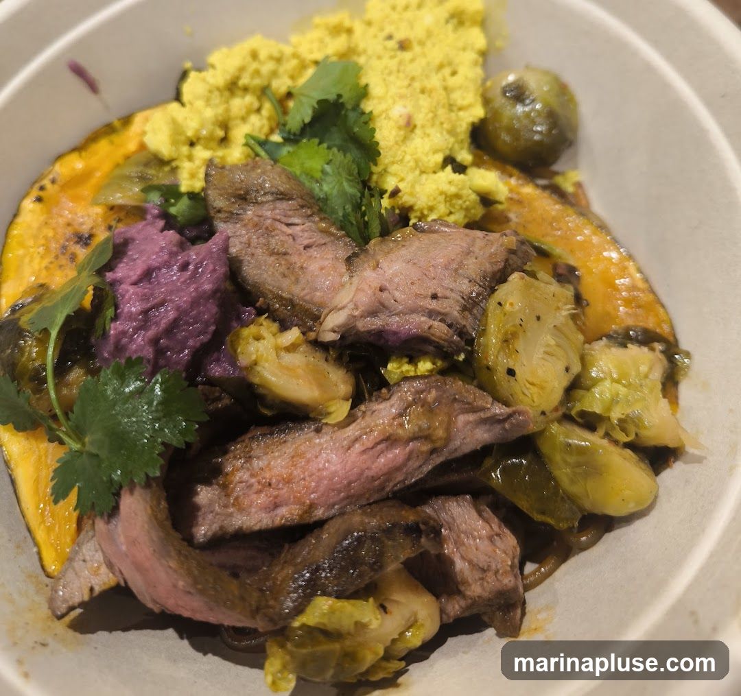 A close-up view of a bowl filled with sliced steak, roasted Brussels sprouts, mashed purple sweet potato, and a yellow crumble, possibly scrambled eggs or corn. The dish is garnished with fresh cilantro and appears to be served in a disposable bowl.