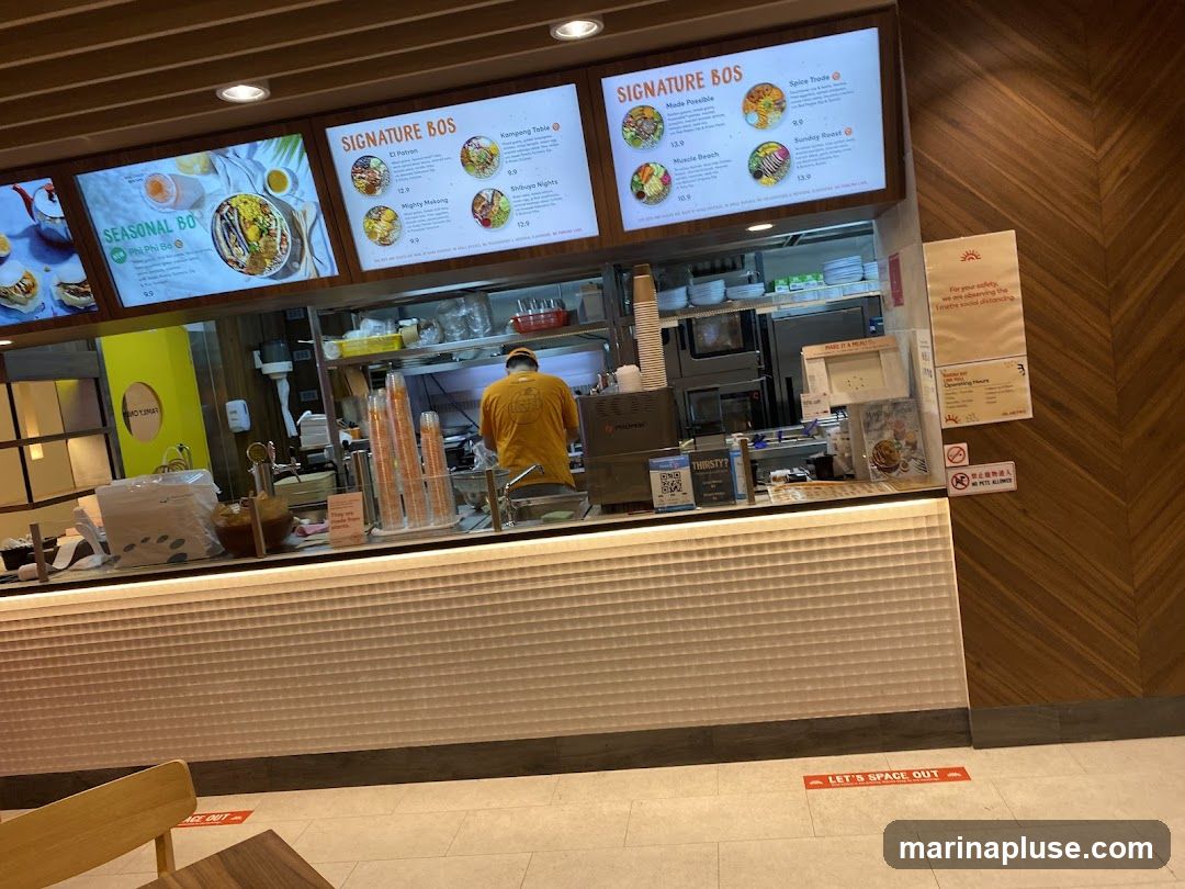 A food stall counter with digital menus displaying seasonal and signature dishes. A staff member in a yellow shirt is visible behind the counter. Social distancing markers are on the floor.