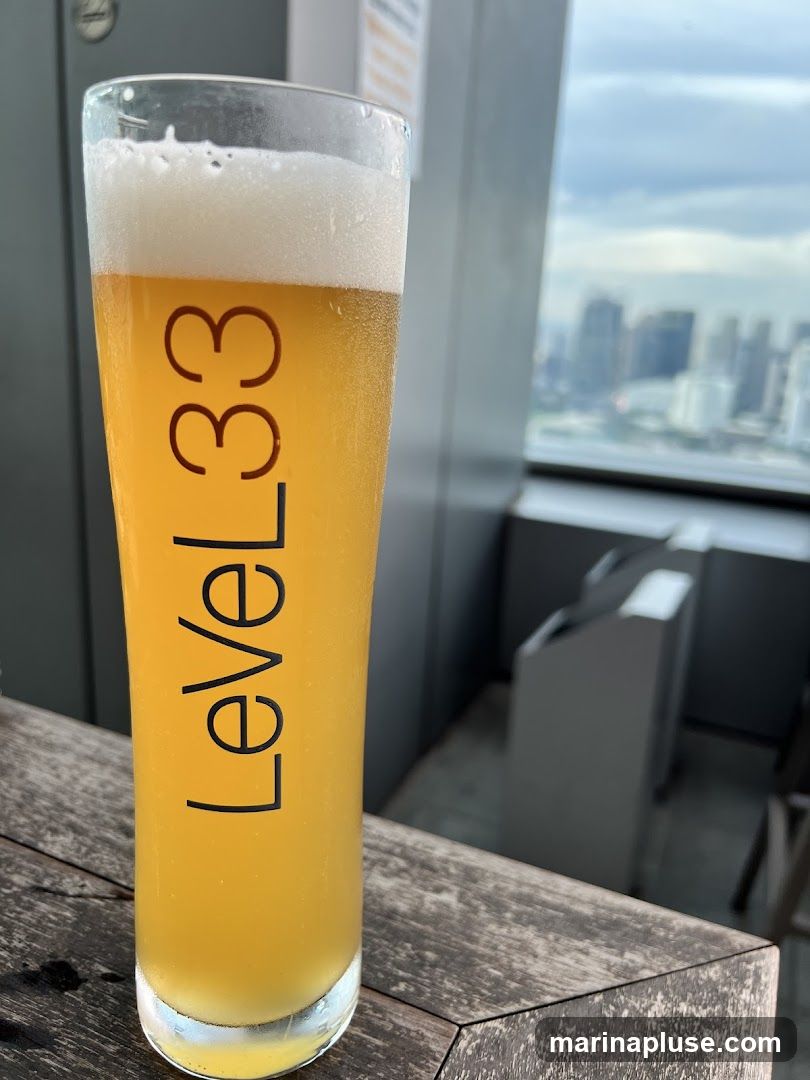 A tall glass of golden beer with a frothy head sits on a wooden table. The glass is branded with the text "Level33" vertically. In the background, a blurred cityscape is visible through a window, suggesting a high-rise location.