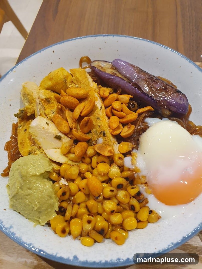 A close-up shot of a noodle bowl filled with various ingredients. The bowl contains noodles, sliced chicken breast seasoned with spices, roasted corn kernels, grilled eggplant slices, a dollop of green paste (possibly avocado or pesto), and a soft-boiled egg with a runny yolk. Peanuts are sprinkled over the chicken and noodles. The dish is served in a light-colored bowl with a blue rim, placed on a wooden surface.