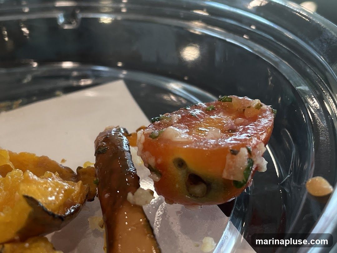 A close-up shot shows a halved cherry tomato topped with chopped herbs and onions, placed in a clear plastic container. Next to the tomato are pieces of what appear to be roasted vegetables and a sausage. The lighting suggests an indoor setting.