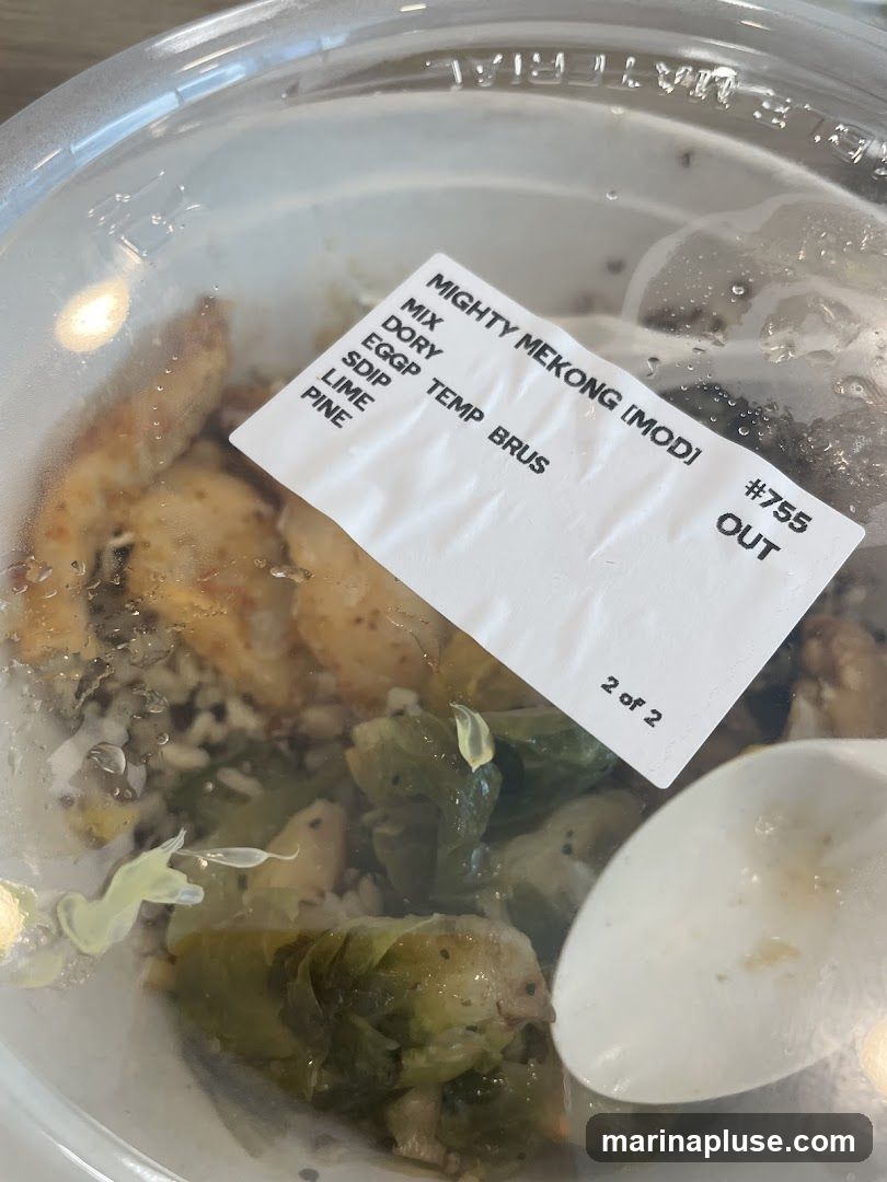 A close-up of a takeout container filled with food, including chicken, Brussels sprouts, and grains. A label on the lid reads "MIGHTY MEKONG [MOD]", order number "#755 OUT", and lists ingredients or components: "MIX", "DORY", "EGGP TEMP BRUS", "SDIP", "LIME", "PINE". The label also indicates "2 of 2".