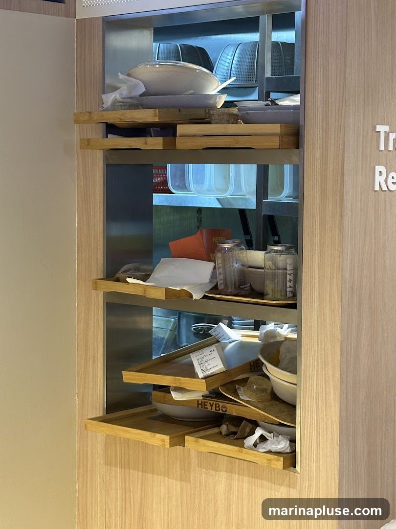 Three wooden shelves are built into a wall, displaying various kitchen items. The top shelf holds large white bowls and what appear to be dish racks. The middle shelf contains plastic containers and jars. The bottom shelf has more bowls, some with the logo "HEYBO" visible, along with receipts and crumpled paper.