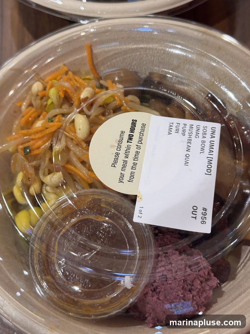 A close-up of a takeout bowl containing a soba noodle dish with various toppings and a side of sauce. The bowl has a label with the text "UNA UMAI [MOD] SOBA BOWL" and a list of ingredients including UNAG, MUSHBEAN QUAI, PURP, FURI, and TAMA. Another label advises to consume the meal within two hours of purchase.