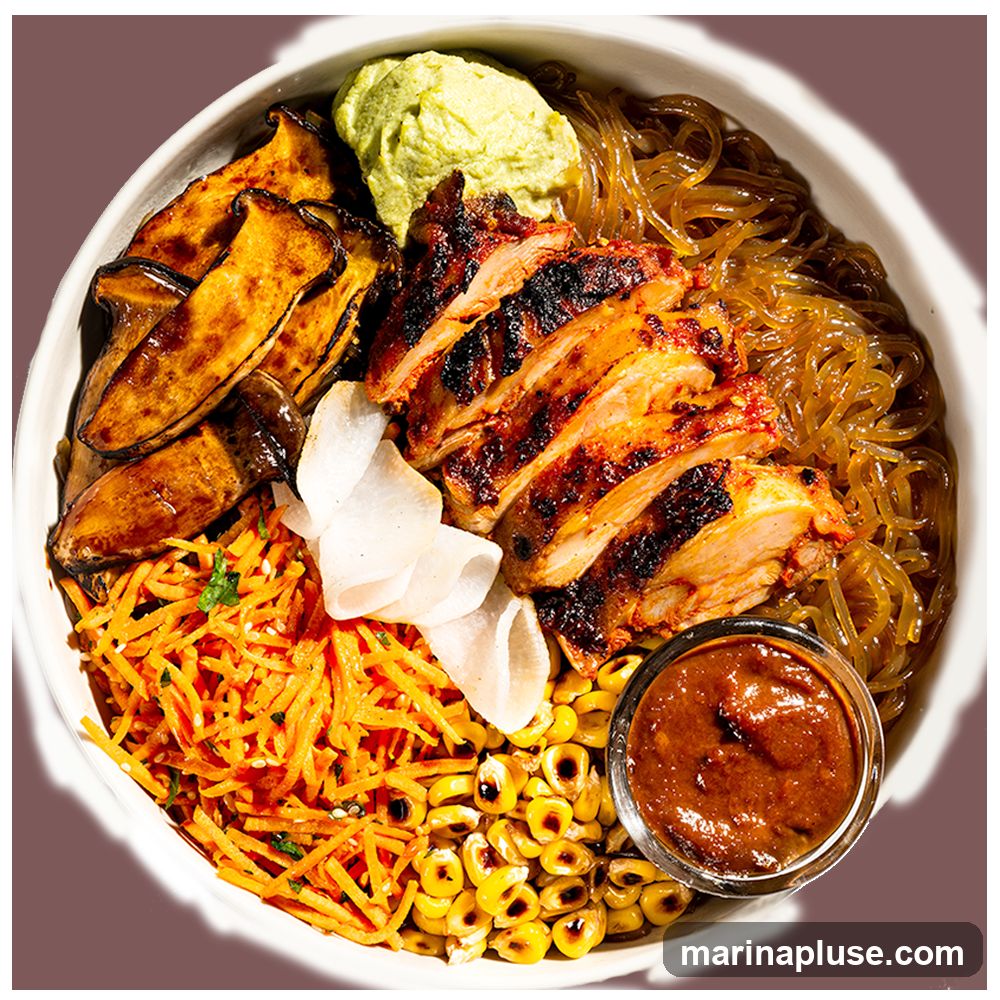 A close-up overhead shot of a chicken bowl. The bowl contains sliced grilled chicken, sweet potato wedges, shredded carrots, corn, glass noodles, a dollop of green sauce, and a small dish of dipping sauce.