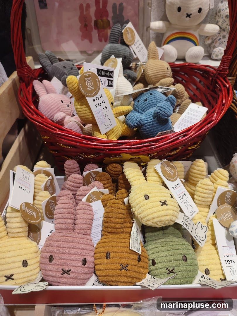 A display of various plush bunny toys in different colors, including pink, yellow, blue, brown, and green. Some toys are in a red basket, while others are arranged in rows on a shelf. Many of the toys have tags attached, some with the brand name "BON TON TOYS" and a logo. In the background, larger plush toys are visible, including one with a rainbow.
