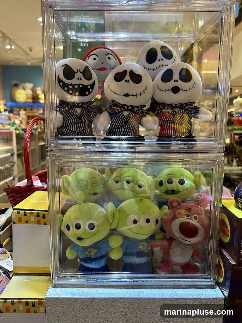 A clear plastic display case filled with plush toys. The top section contains several Jack Skellington plush toys and one Sally plush toy from The Nightmare Before Christmas. The bottom section contains several green alien plush toys from Toy Story and one Lotso Huggin' Bear plush toy from Toy Story 3. The display is on a shelf in what appears to be a store, with other merchandise visible in the background.