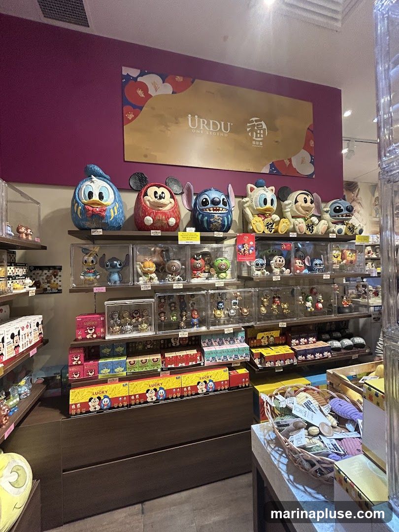 A retail display featuring various Disney characters, including Donald Duck, Mickey Mouse, Stitch, and others, presented as collectible figures and plush toys. The merchandise is arranged on shelves and in display cases, with some items packaged in boxes labeled "Lucky." A large banner with the text "URDU ONE LEGEND" and Chinese characters is visible on the wall above the display. A basket filled with small items is on a counter in the foreground.