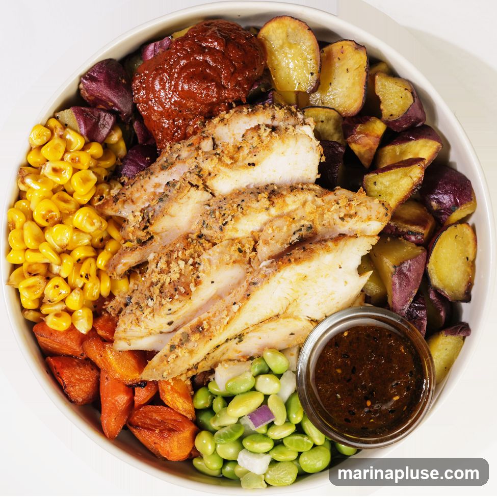 A close-up overhead view of a bowl filled with food. The bowl contains sliced, seasoned chicken breast, corn, roasted sweet potatoes, roasted carrots, edamame with diced red onion, and a dollop of red sauce. A small glass dish of dark sauce is also visible.