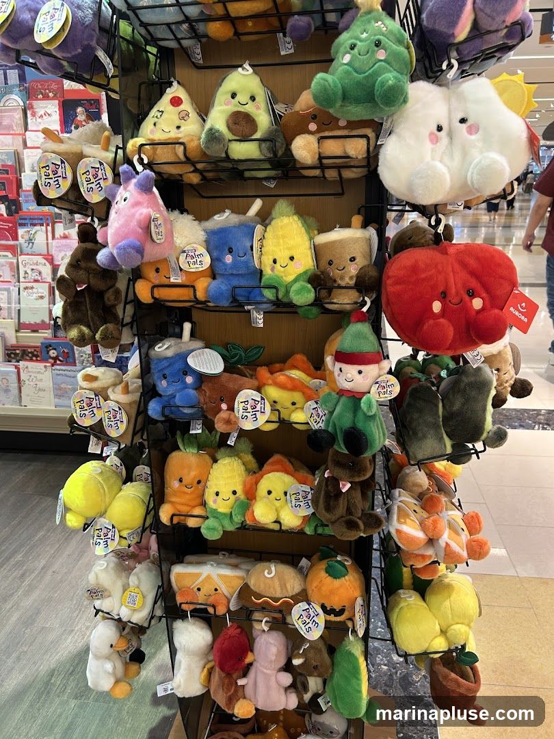 A display rack filled with various plush toys, including food items like pizza, avocado, corn, and drinks, as well as other characters like clouds and a red apple. Some toys have tags that say "Palm Pals" and "Aurora". In the background, there are greeting cards and blurred figures of people shopping.