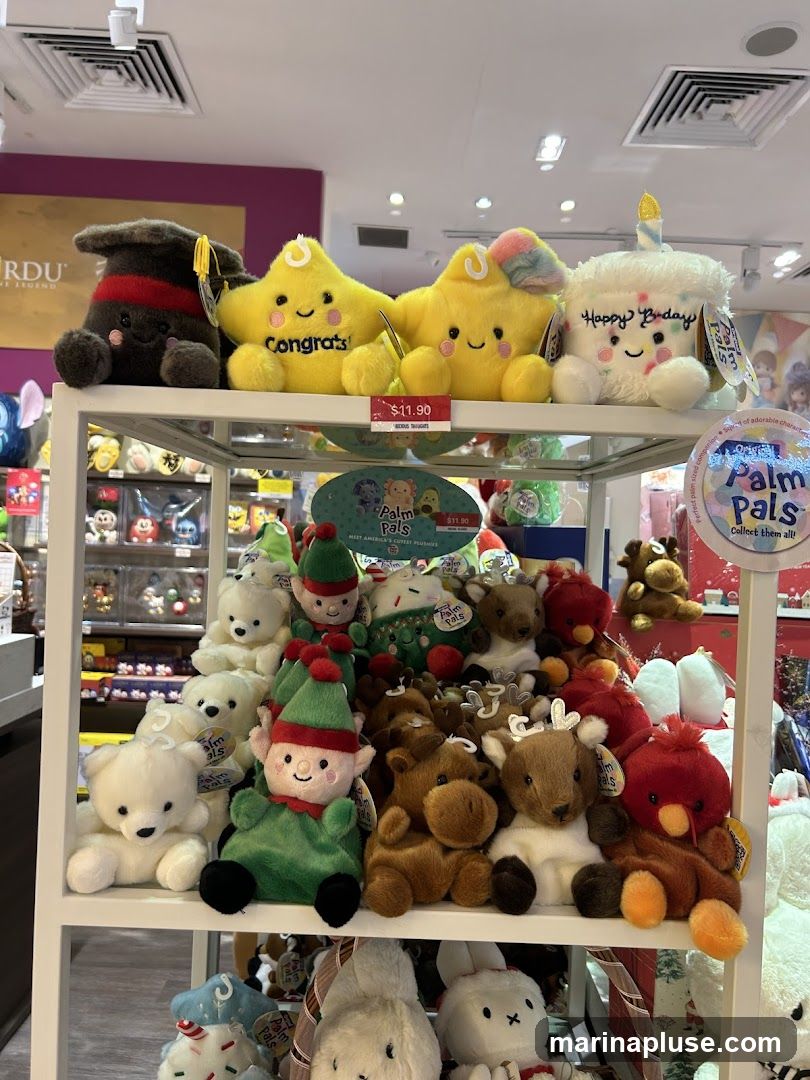 A display shelf filled with various plush toys, including graduation caps, smiling stars, a birthday cake, elves, polar bears, and reindeer. Some toys have price tags indicating $11.90. The background shows more merchandise and store shelving.