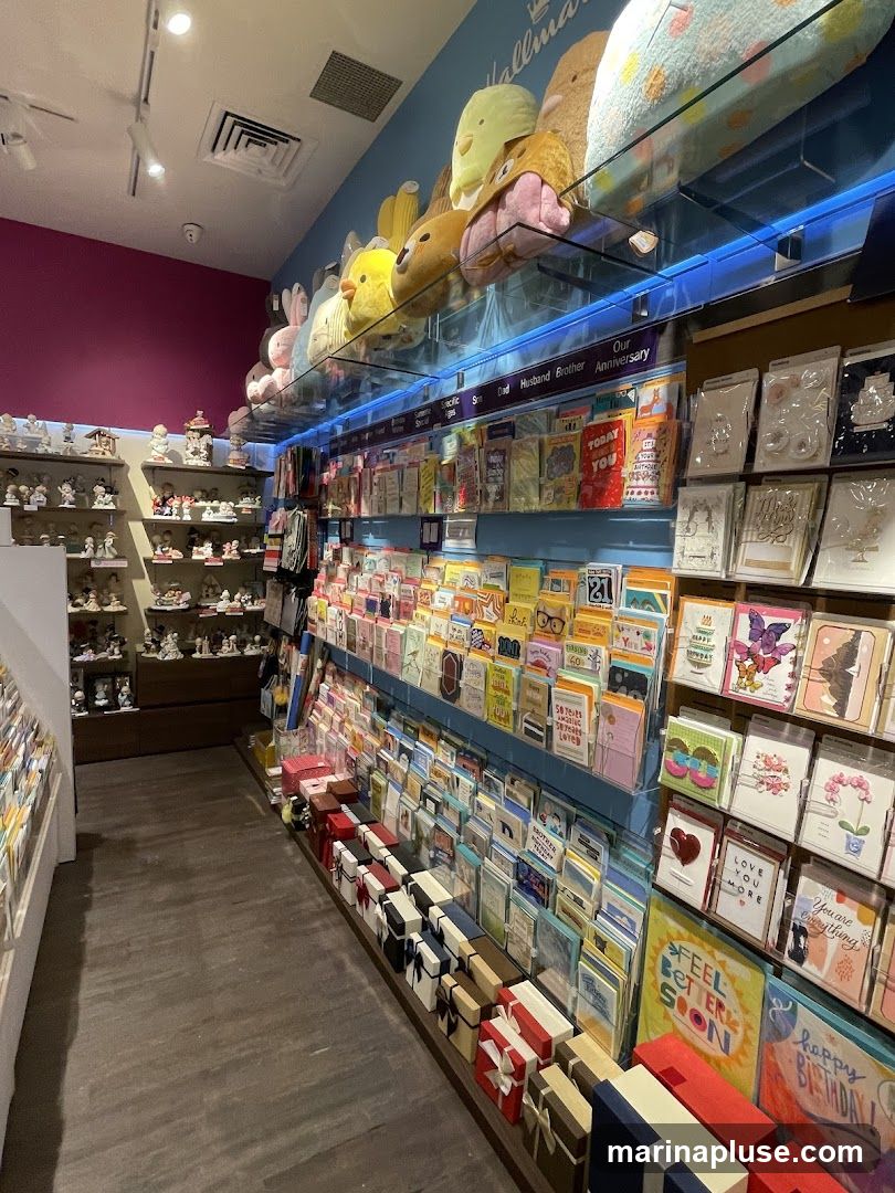 The interior of a Hallmark store, showcasing a wide selection of greeting cards, gift boxes, and plush toys. The shelves are stocked with various cards categorized by occasion, such as birthdays, anniversaries, and general greetings. Gift boxes are neatly arranged on the floor and shelves. Plush toys are displayed on a shelf above the cards. The store has a bright and organized appearance.