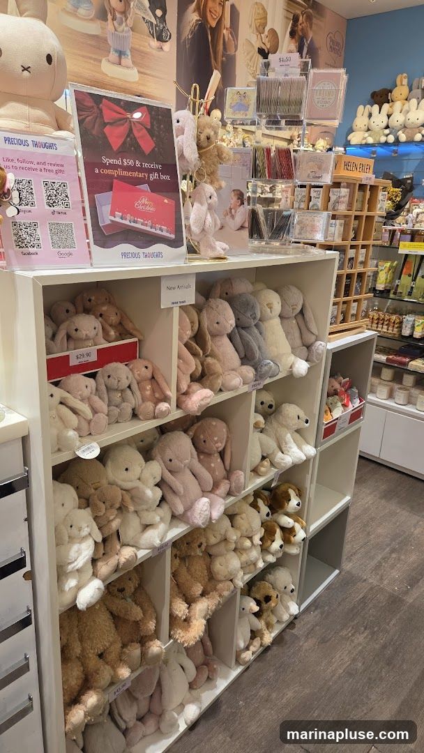 A display of plush toys, primarily bunnies and teddy bears, arranged on shelves. A sign advertises a promotion: "Spend $50 & receive a complimentary gift box." Other items visible include greeting cards and decorative figures.