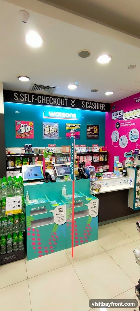 The image shows the checkout area of a Watsons store. There are self-checkout stations and a cashier counter. Promotional signs for discounts are displayed on the wall. Recycling bins are placed in front of the checkout counters. Shelves stocked with various products are visible in the background.