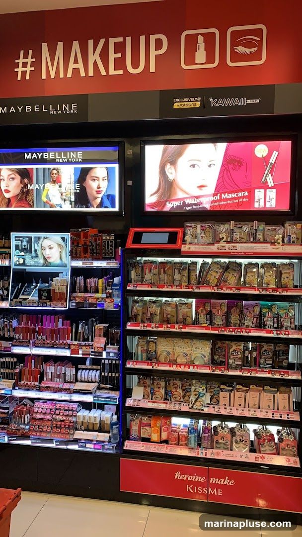 A retail display featuring various makeup products, primarily from Maybelline New York. The display includes shelves stocked with lipsticks, mascaras, eyeshadows, and other cosmetics. Digital screens showcase advertisements and product images. The overall aesthetic is bright and commercial, with prominent branding.