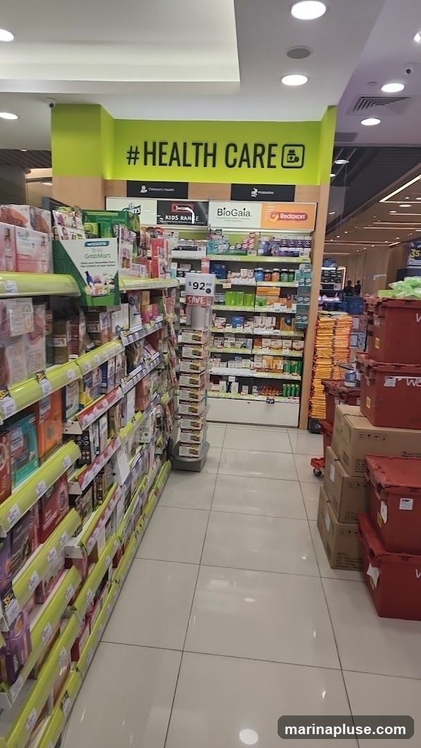 A view down an aisle in a pharmacy or health store, with shelves stocked with various health and personal care products. A prominent green sign above reads "#HEALTH CARE" with a medical cross icon. Products are displayed on shelves on both sides of the aisle, with some promotional signage visible. Stacks of boxes and red storage containers are seen further down the aisle.