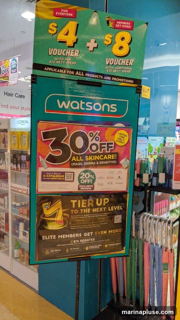 A display board shows various promotions for Watsons. The top section offers $4 and $8 vouchers for everyone and members respectively, with minimum nett spend. Below that, a prominent "30% OFF ALL SKINCARE!" offer is advertised, with an additional "20% OFF! EXCLUSIVE BRANDS" section. The bottom part promotes a "TIER UP TO THE NEXT LEVEL" program for elite members, offering $15 rebates and other benefits. The display also shows umbrellas for sale.