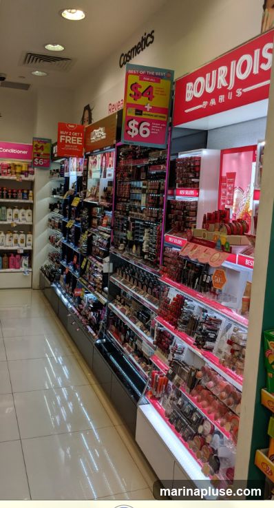 A view of a cosmetics store aisle with shelves stocked with various makeup and beauty products. Promotional signs are visible, including one advertising "BUY 2 GET 1 FREE" and another with prices of $4 and $6. The Bourjois Paris brand is prominently displayed.