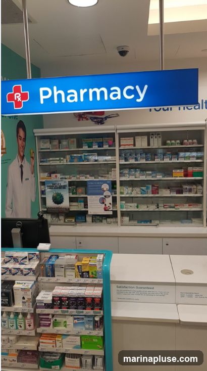 A view inside a pharmacy with shelves stocked with various medications and health products. A prominent blue sign with a red cross and the word "Pharmacy" is visible overhead. To the left, a poster features a man in a white coat holding an apple.