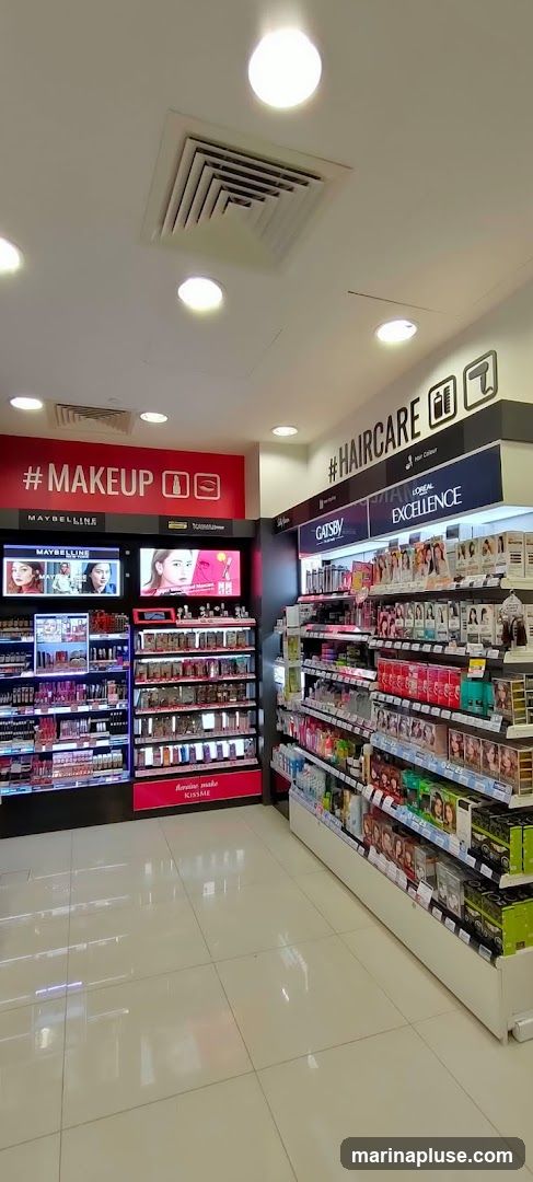A well-lit interior view of a retail store showcasing makeup and haircare products. The left side features a prominent red display with "# MAKEUP" and Maybelline branding, displaying various makeup items. The right side has a black and white sign with "# HAIRCARE" and icons for hair dryer and comb, with shelves stocked with L'Oreal Excellence hair color and Gatsby products. The floor is tiled and reflective.