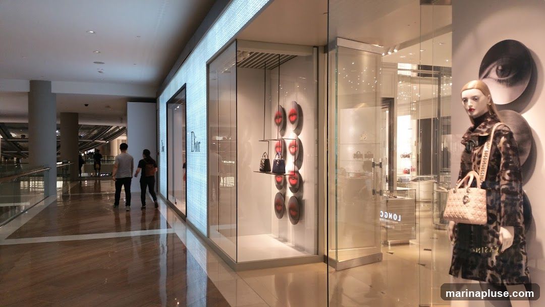 A well-lit interior view of a Dior store, showcasing its merchandise. The storefront features a large, illuminated facade with the Dior logo. Inside, a mannequin dressed in a leopard-print coat displays a handbag. The display window also features a collection of handbags on shelves, with decorative circular images of lips on the wall behind them. The polished floor reflects the store's interior and the surrounding mall.