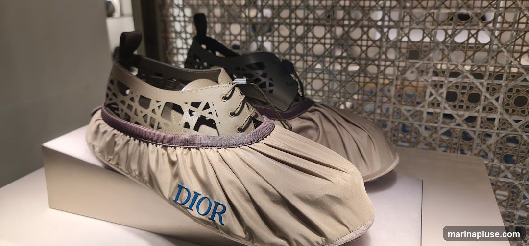 A close-up shot of two pairs of Dior shoes with a distinctive woven pattern on the upper and a gathered fabric overlay on the toe. The Dior logo is visible in blue on the overlay of the shoe in the foreground. The shoes are displayed on a white surface against a backdrop of intricate woven material.