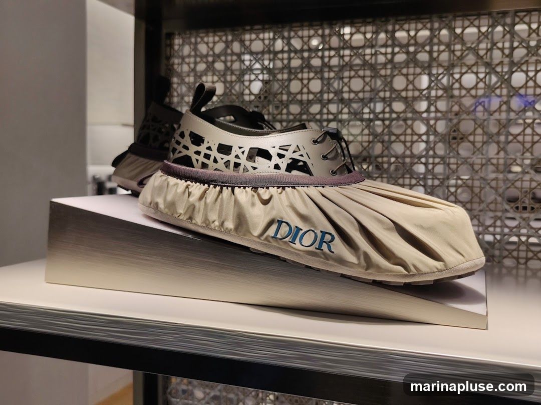 A pair of Dior sneakers with a unique pleated fabric overlay and cut-out detailing on the upper. The Dior logo is visible in blue on the overlay. The shoes are displayed on a slanted white stand against a backdrop of a woven pattern.