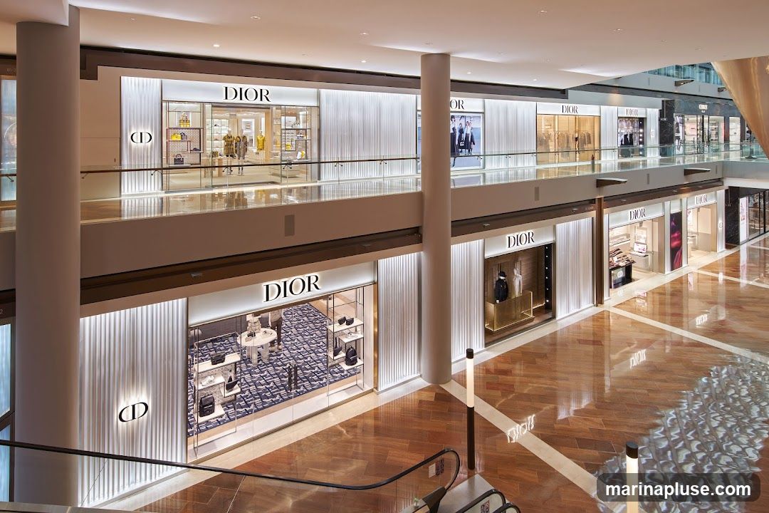 A wide shot of multiple Dior boutiques on two levels of a modern, well-lit shopping mall. The boutiques feature clean, minimalist designs with illuminated signage and displays showcasing handbags, clothing, and accessories. The mall has polished wooden floors and escalators visible in the foreground.