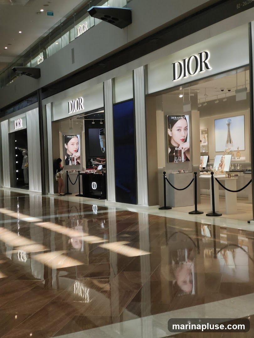 The exterior of a Dior store with large illuminated signs displaying the brand name. The storefront features large windows showcasing displays of makeup and perfume. A poster of a woman wearing makeup is visible in one of the windows. The floor in front of the store is highly polished and reflects the store's facade and overhead lighting.