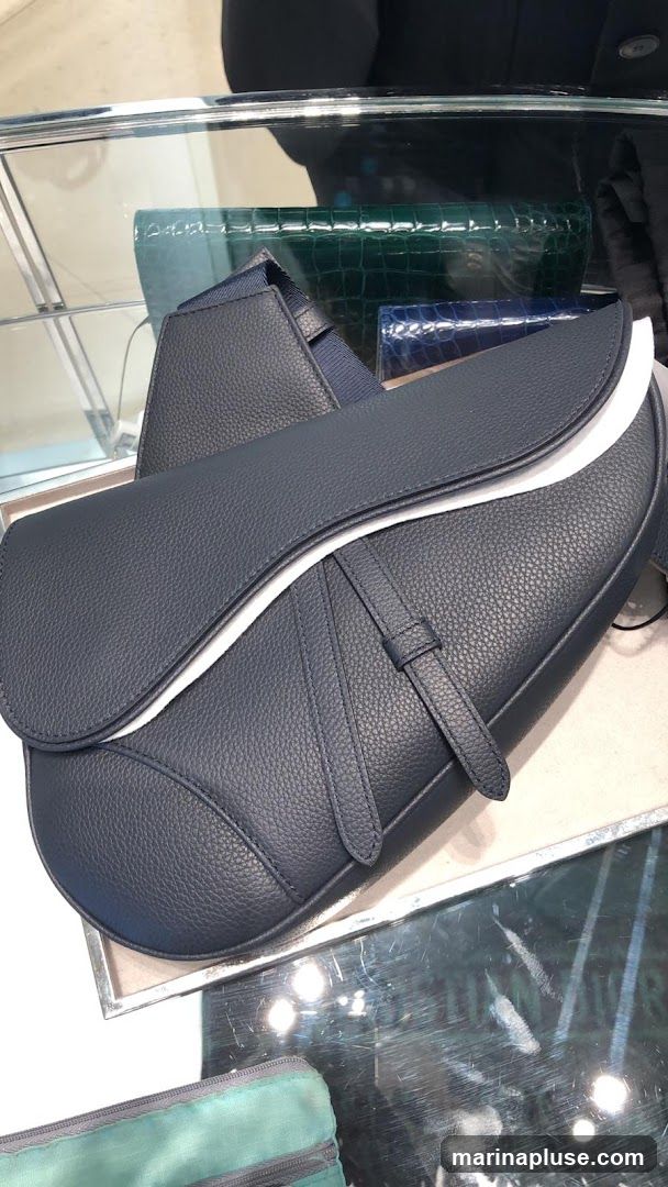 A close-up view of a dark blue leather saddle bag with white trim and a blue strap. The bag is displayed on a reflective surface, possibly a glass counter, with other accessories visible in the background, including a green crocodile-embossed clutch and a blue patterned clutch.