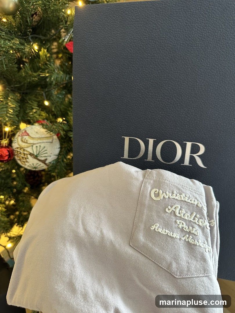 A dark blue Dior gift box is placed behind a light-colored garment with embroidered text. The text on the garment reads "Christian Dior Atelier Paris Avenue Montaigne". A decorated Christmas tree with lights and ornaments is visible to the left of the box.