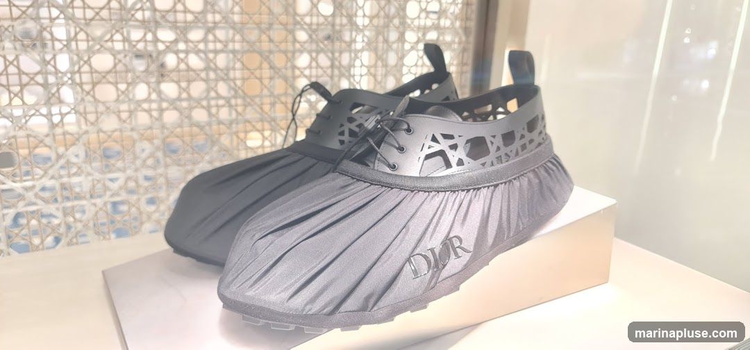 A pair of black Dior shoes with a pleated fabric overlay and a cut-out pattern on the upper. The shoes are displayed on a white surface with a light-colored, woven background. The Dior logo is visible on the side of one shoe.