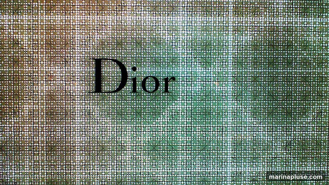 The image features the Dior logo in black text against a textured background. The background has a grid pattern with small circular shapes within each square of the grid. The colors of the background transition from a brownish-green on the left to a greener hue on the right.