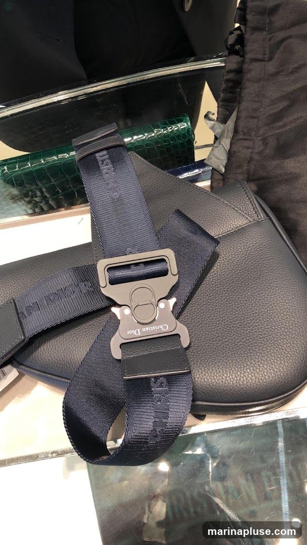 A close-up view of a dark blue Christian Dior saddle bag with a prominent buckle and strap. The strap features the "Christian Dior" logo woven into the fabric. A green crocodile-embossed clutch is visible behind the bag.
