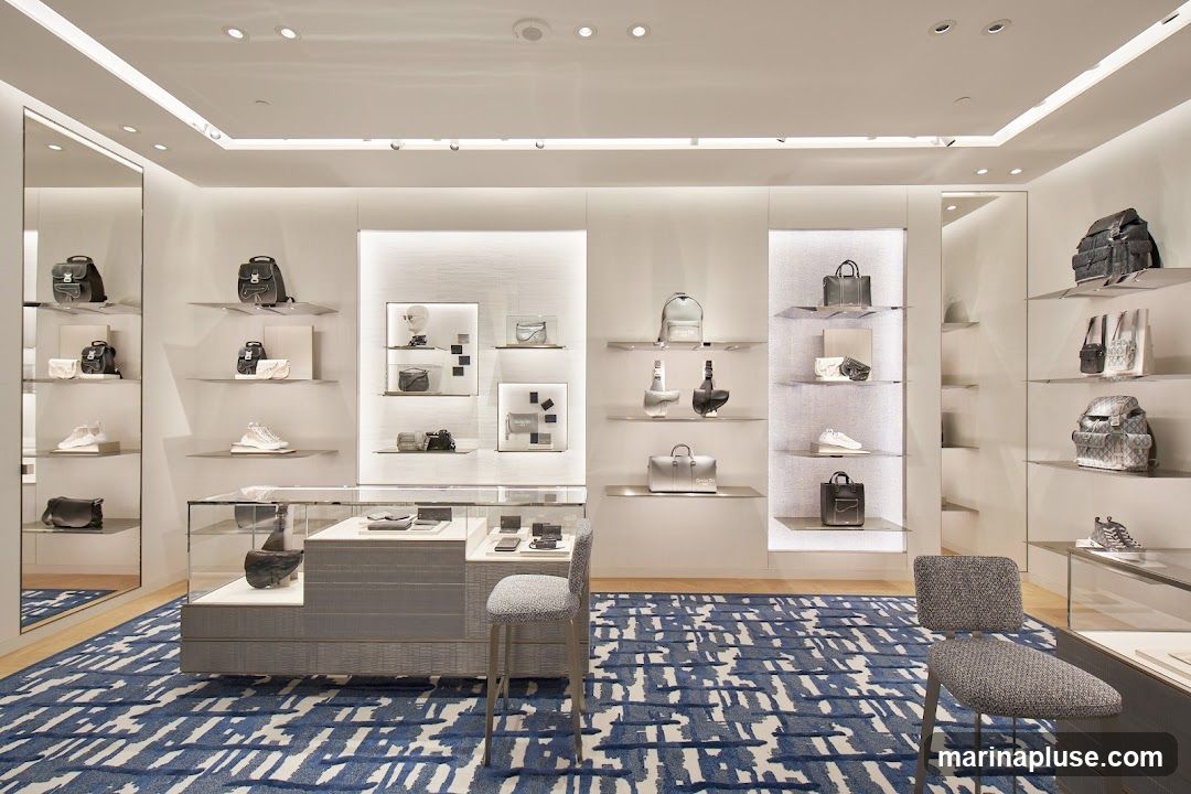 A well-lit, modern retail space displaying a collection of luxury handbags, backpacks, and shoes on shelves and in display cases. The store features a distinctive blue and white patterned carpet and minimalist decor. Two grey upholstered stools are placed in the foreground.