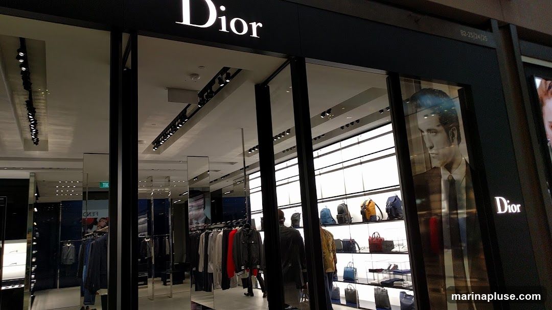 The exterior of a Dior store with the brand name prominently displayed above the entrance. The storefront features large glass windows showcasing clothing racks with apparel and shelves displaying handbags and backpacks. A large advertisement featuring a man in a suit is visible in one of the windows.