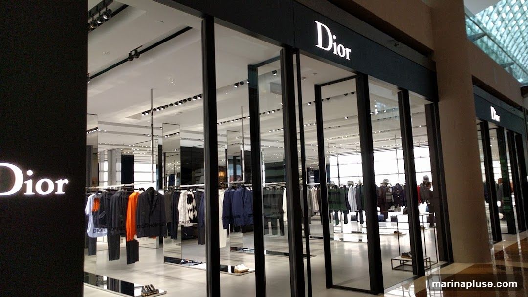 A well-lit, modern storefront for the luxury brand Dior. The facade features large glass windows and doors, allowing a view into the store's interior. The Dior logo is prominently displayed in white lettering on a dark background above the entrance. Racks of men's clothing, including jackets, shirts, and trousers, are visible inside, along with display shelves showcasing shoes. The store appears to be located within a shopping mall, with a ceiling featuring a grid-like structure and ample lighting.