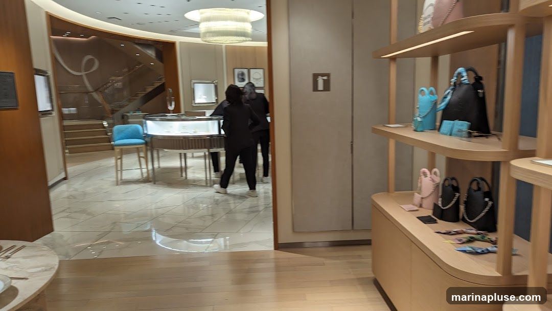 The image shows the interior of a high-end retail store, likely a jewelry or handbag boutique. In the foreground, shelves display various handbags in colors like black, pink, and blue. Further inside, a staircase leads to an upper level. Display cases with jewelry are visible, and two staff members in dark uniforms are attending to customers or displays. The store features a modern and elegant design with marble flooring and warm wood accents.