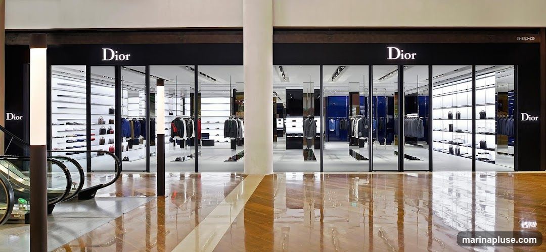A wide shot of a Dior store's facade in a mall. The storefront features large glass windows displaying clothing and handbags on shelves. The Dior logo is prominently displayed in white lettering on a black background above the windows. An escalator is visible to the left of the store entrance. The floor is polished and reflects the store's interior and exterior.