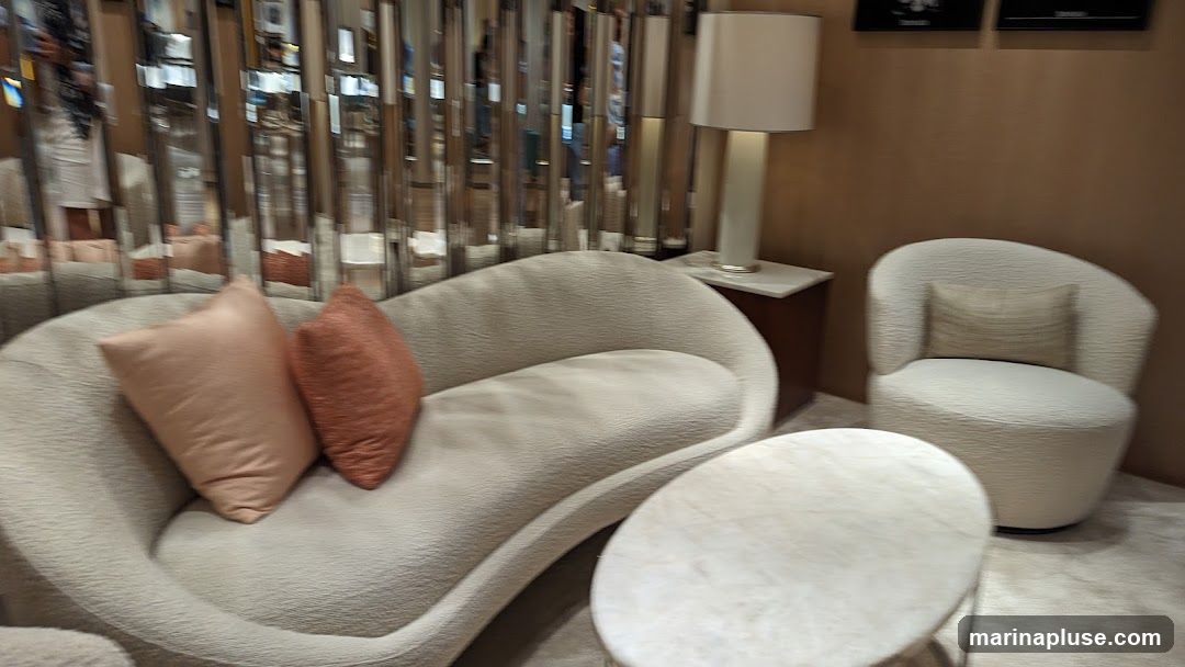 A display of modern living room furniture, including a curved sofa with two accent pillows, a round coffee table, and a plush armchair. The background features a wall with vertical mirrored panels and a table lamp on a side table.
