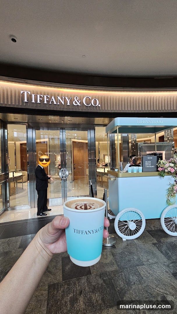 A hand holds a Tiffany & Co. branded coffee cup with latte art in front of a Tiffany & Co. store. A light blue coffee cart with the Tiffany & Co. logo is parked to the right of the store entrance. The cart has a menu board displaying "BLACK WHITE MOCHA CHOCOLATE".