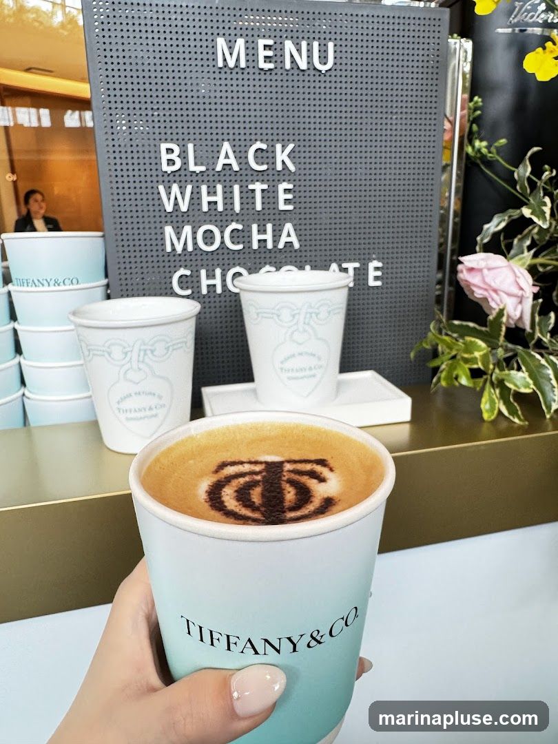 A hand holds a light blue and white paper cup with the Tiffany & Co. logo and a latte art design on top of the coffee. In the background, a dark gray pegboard menu displays