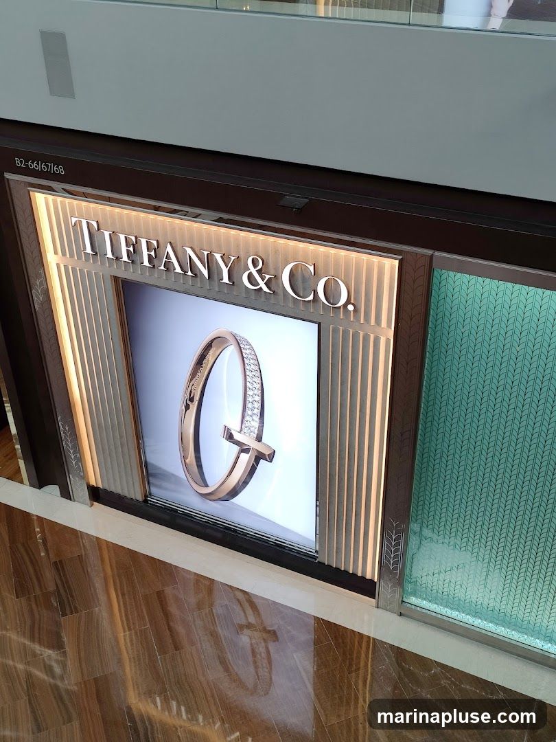 A display window for Tiffany & Co. features a large illuminated advertisement of a rose gold bracelet with diamonds. The Tiffany & Co. logo is prominently displayed above the advertisement. To the right of the display is a section of turquoise glass with a textured pattern. The floor in front of the display is made of polished brown and beige marble tiles.